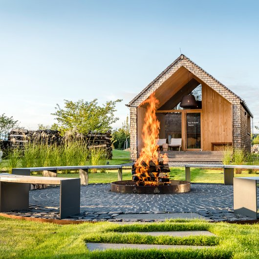 Seezeit re.connect – Bewusste Verbindung mit Natur & Kraftort Offene Feuerstelle mit hölzernen Bänken vor einer kleinen Holzsauna im Garten