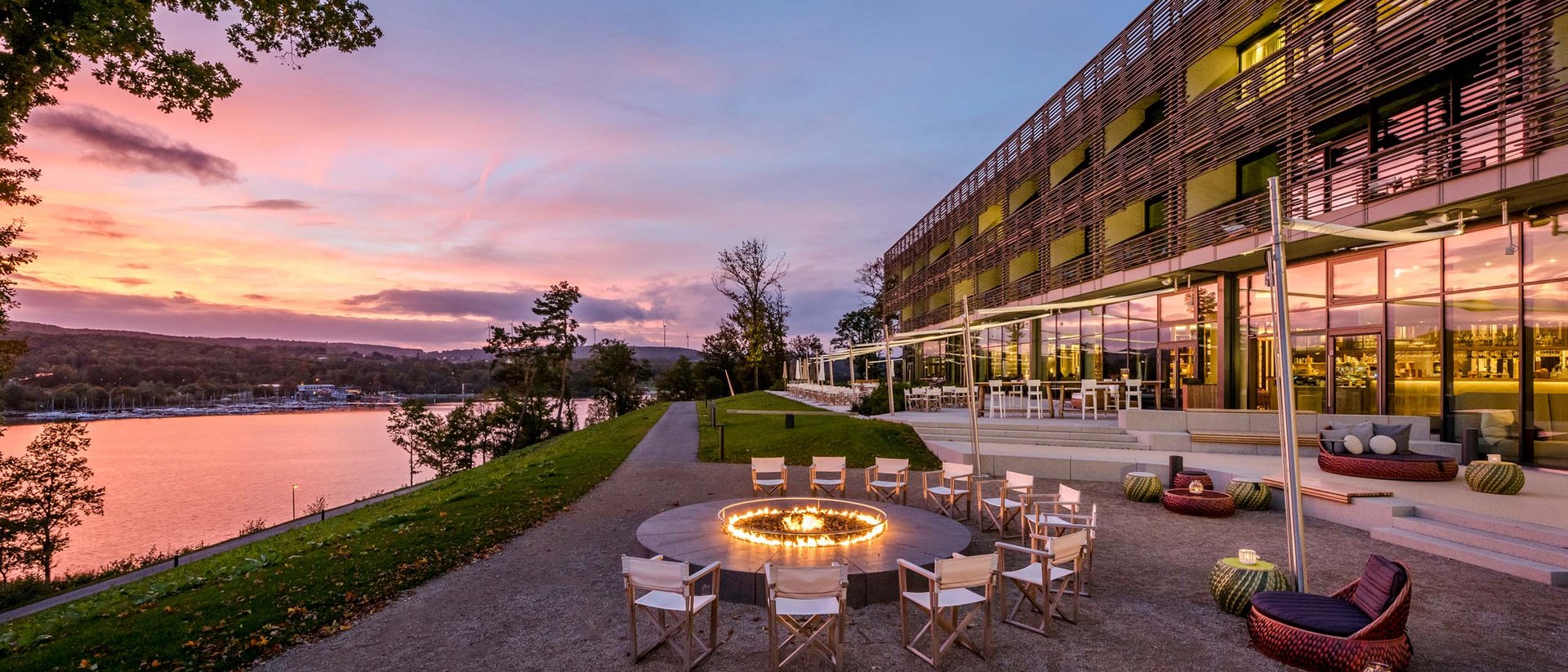 Seezeitlodge : Galerie photos Espace assis avec feu circulaire près d'un bâtiment moderne au bord de la rivière au coucher du soleil