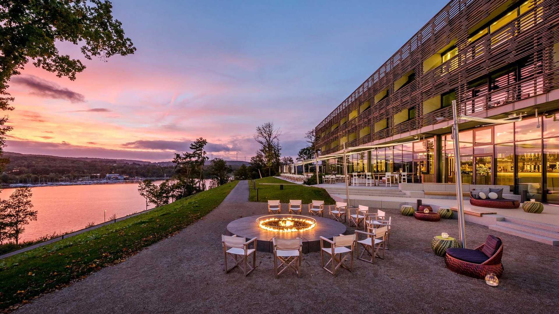 Seezeitlodge: Bildergalerie Sitzbereich mit Feuerring neben modernem Gebäude und Fluss bei Sonnenuntergang