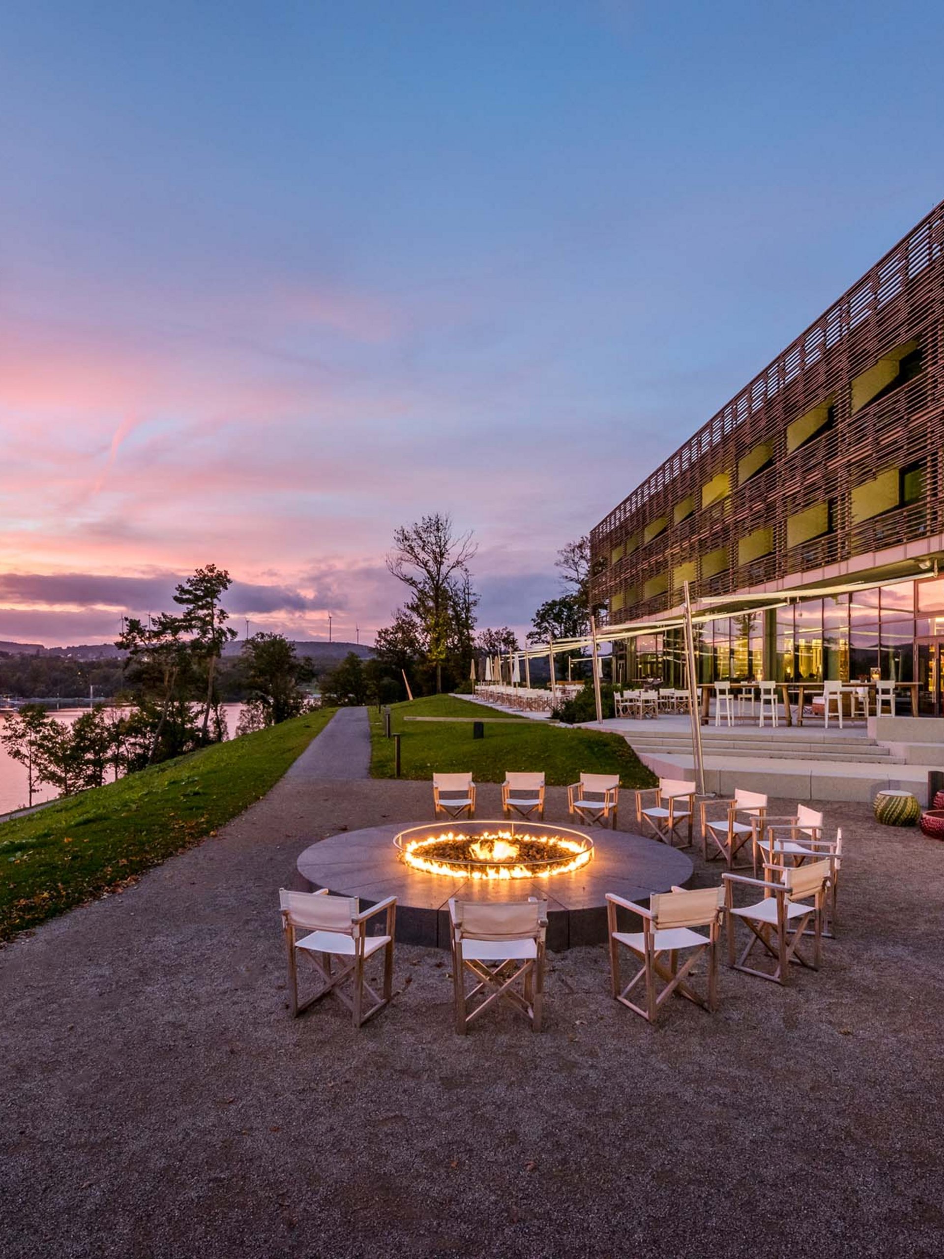 Seezeitlodge : Galerie photos Espace assis avec feu circulaire près d'un bâtiment moderne au bord de la rivière au coucher du soleil