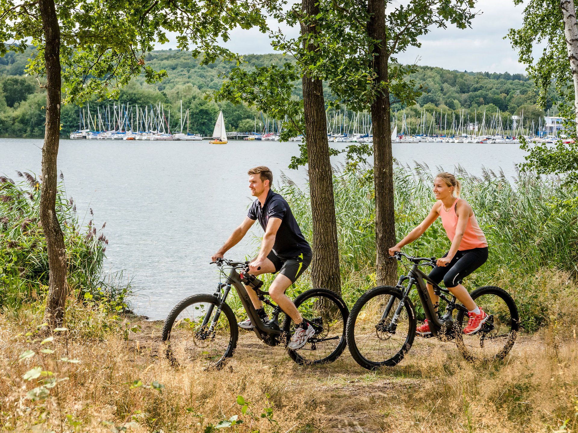 Urlaub am Bostalsee: die Highlights der Region Paar fährt Mountainbikes am Seeufer mit Segelbooten im Hintergrund