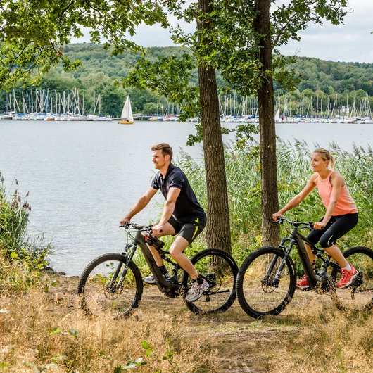 Luxushotel Seezeitlodge: Lassen Sie sich begeistern! Paar fährt Mountainbikes am Seeufer mit Segelbooten im Hintergrund