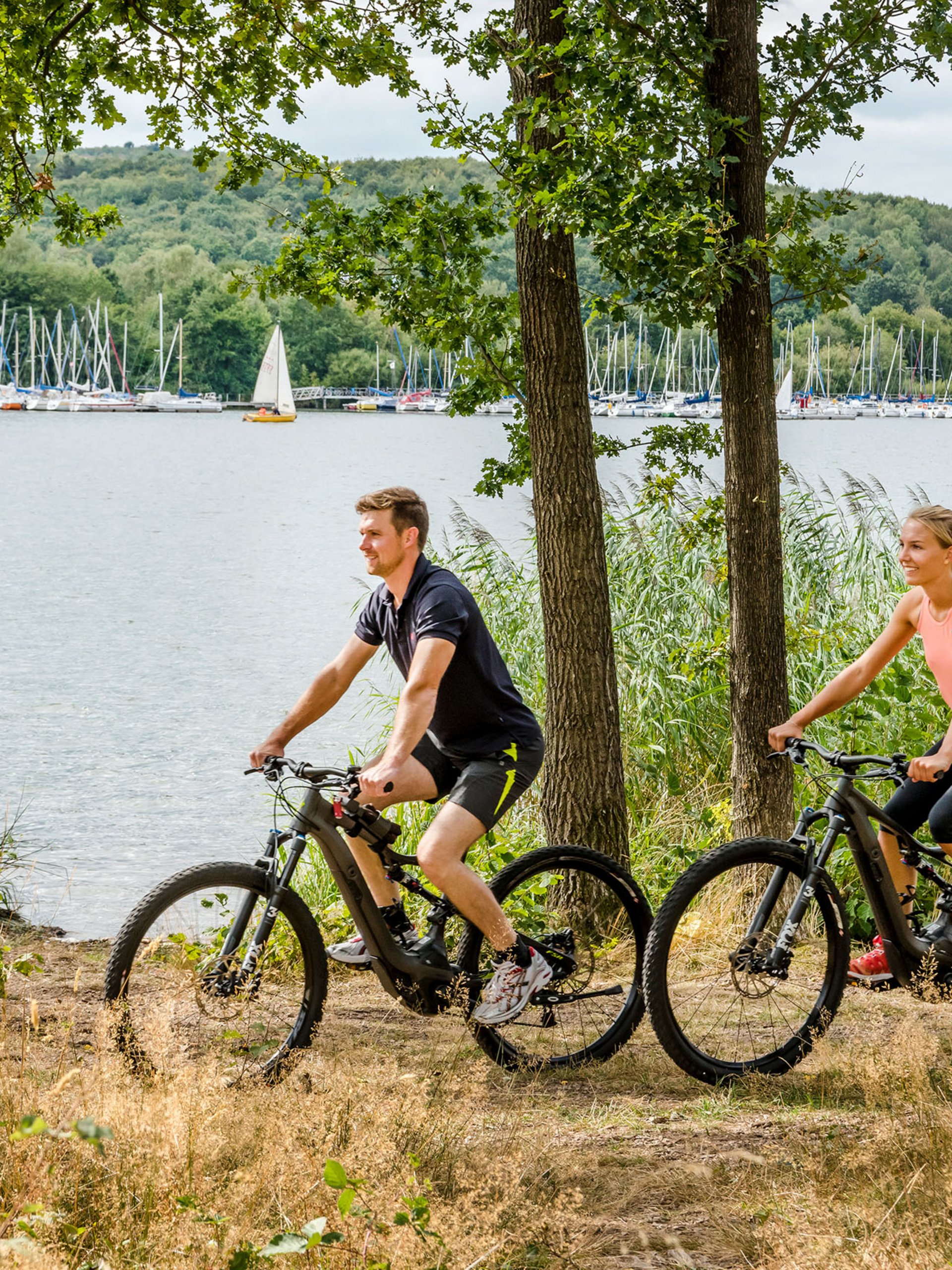 Seezeitlodge: Bildergalerie Paar fährt Mountainbikes am Seeufer mit Segelbooten im Hintergrund