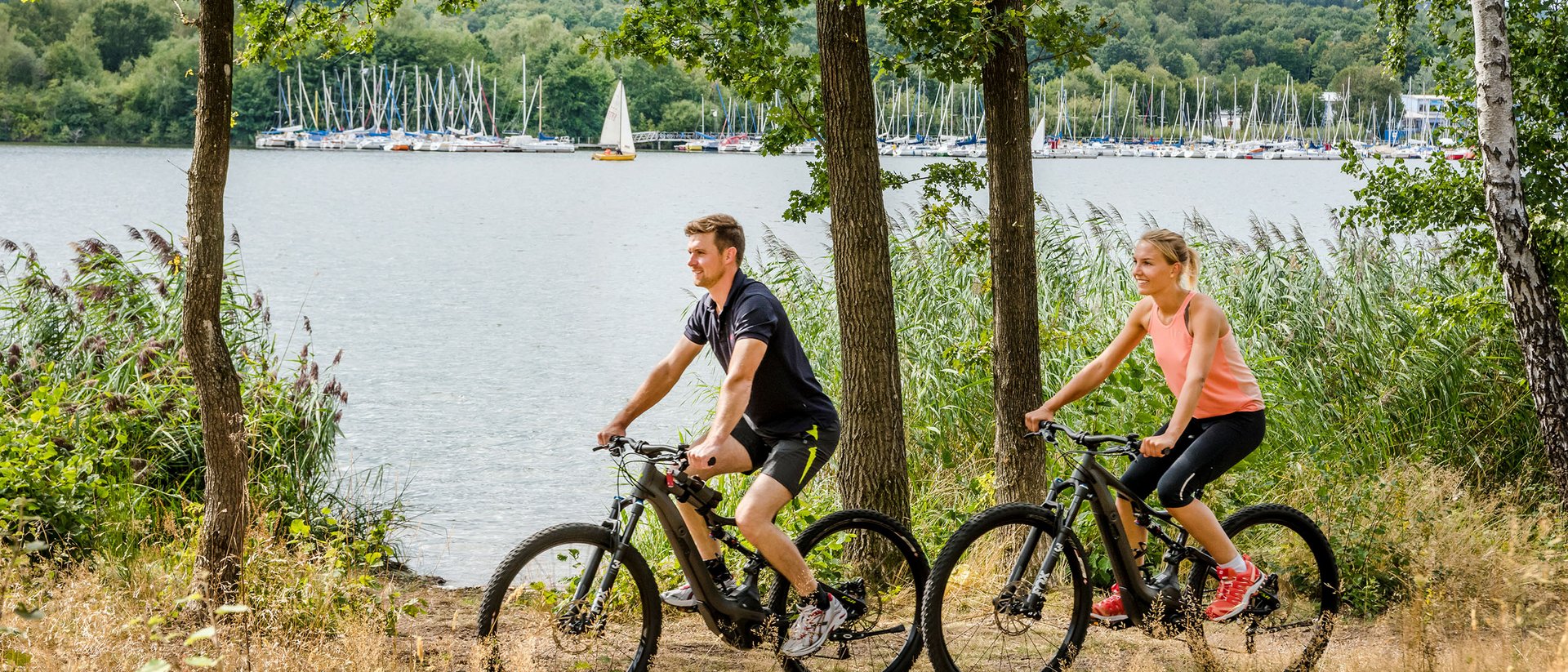 Seezeitlodge: Bildergalerie Paar fährt Mountainbikes am Seeufer mit Segelbooten im Hintergrund