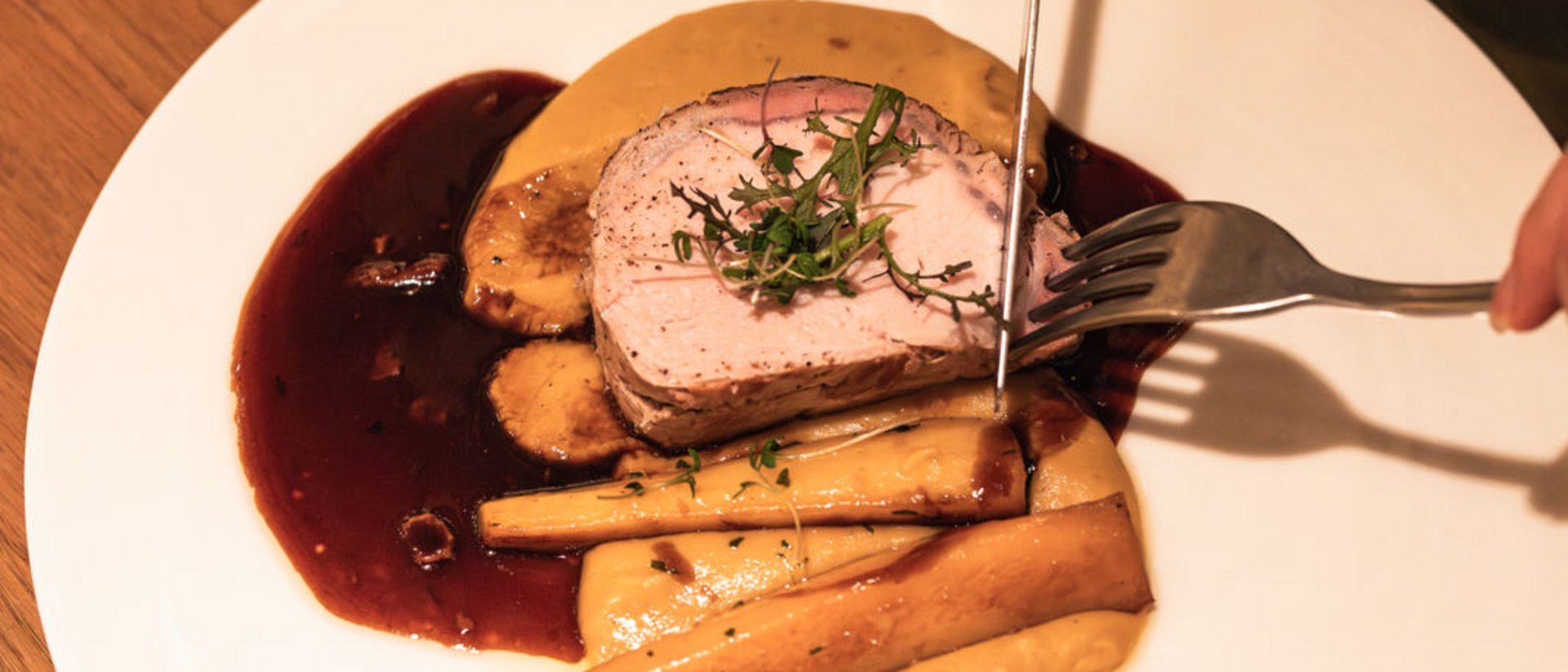 Seezeitlodge: Bildergalerie Person schneidet gebratenes Fleisch mit Beilagen und Soße auf weißem Teller