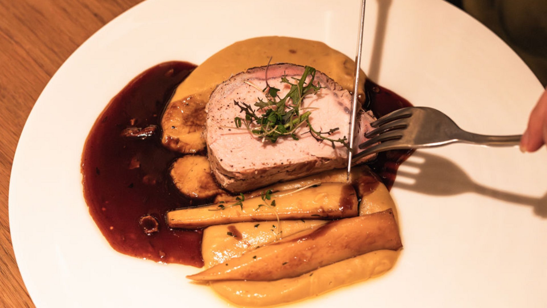 Seezeitlodge: Bildergalerie Person schneidet gebratenes Fleisch mit Beilagen und Soße auf weißem Teller