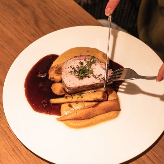 Luxushotel Seezeitlodge: Lassen Sie sich begeistern! Person schneidet gebratenes Fleisch mit Beilagen und Soße auf weißem Teller