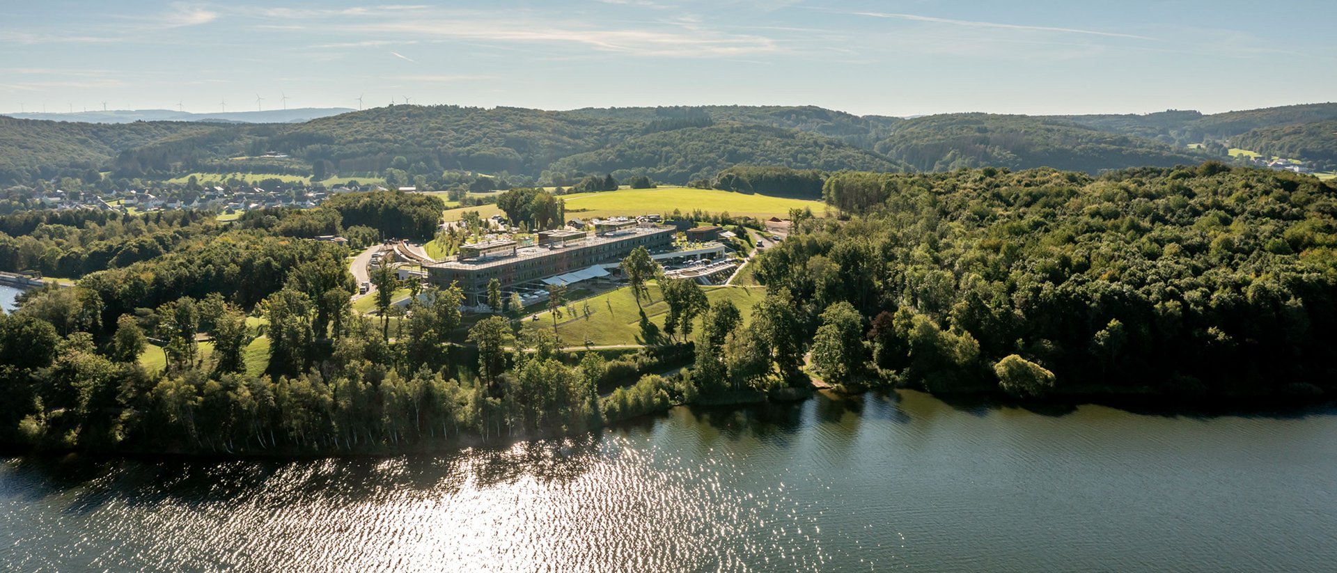 Seezeitlodge: Bildergalerie Luftaufnahme eines Sees mit bewaldeten Ufern und einem großen Gebäude bei Sonnenschein