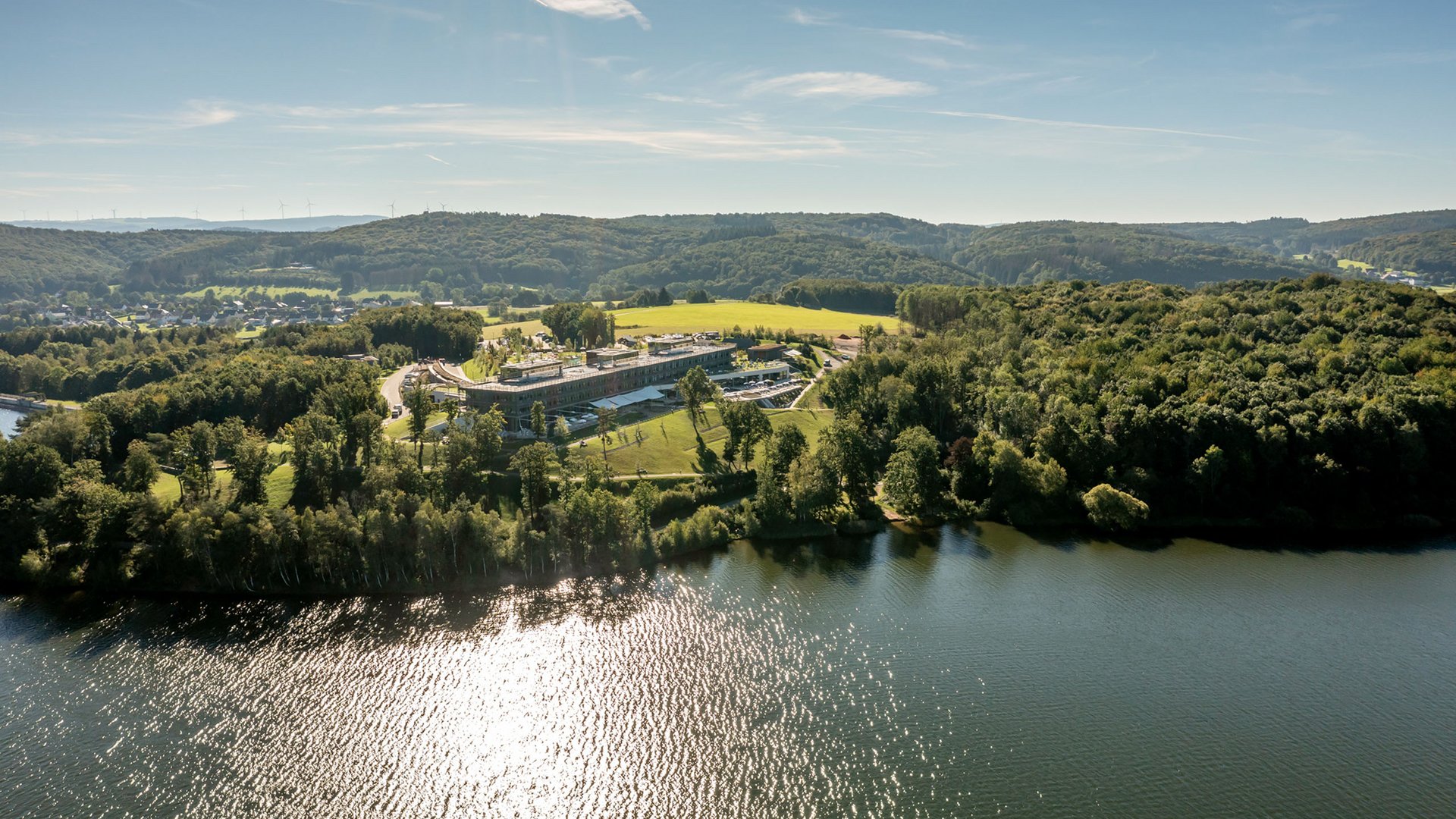 Seezeitlodge: Lieblingsgeschichten Luftaufnahme eines Sees mit bewaldeten Ufern und einem großen Gebäude bei Sonnenschein
