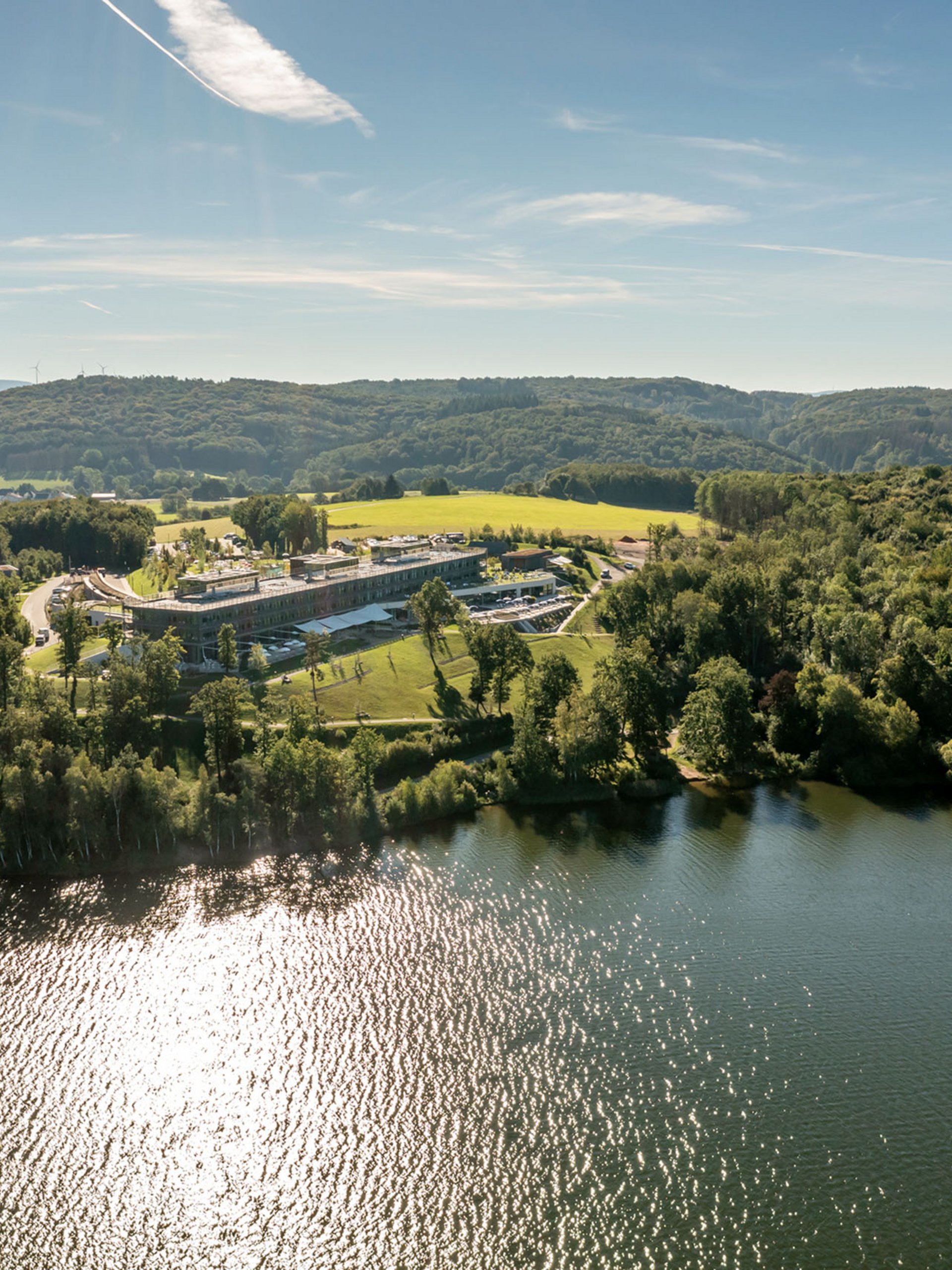 Seezeitlodge: Bildergalerie Luftaufnahme eines Sees mit bewaldeten Ufern und einem großen Gebäude bei Sonnenschein