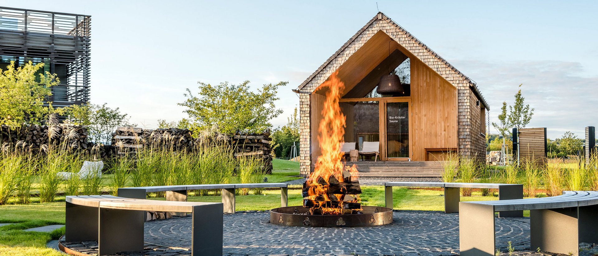 Seezeitlodge: Bildergalerie Feuerstelle mit brennendem Holz und Rundbank vor einer Sauna aus Holz