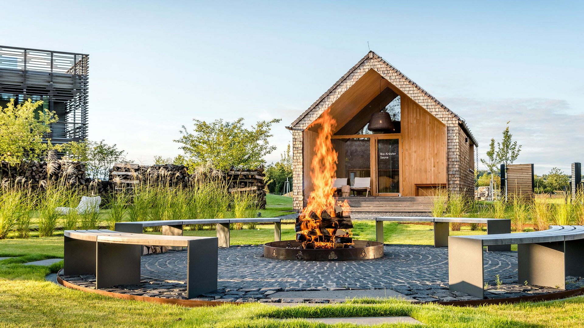Seezeitlodge: Bildergalerie Feuerstelle mit brennendem Holz und Rundbank vor einer Sauna aus Holz