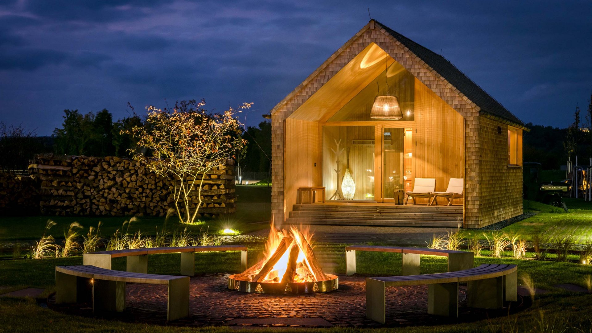 Seezeitlodge : Galerie photos Maison en bois de nuit avec feu de camp et bancs dans le jardin
