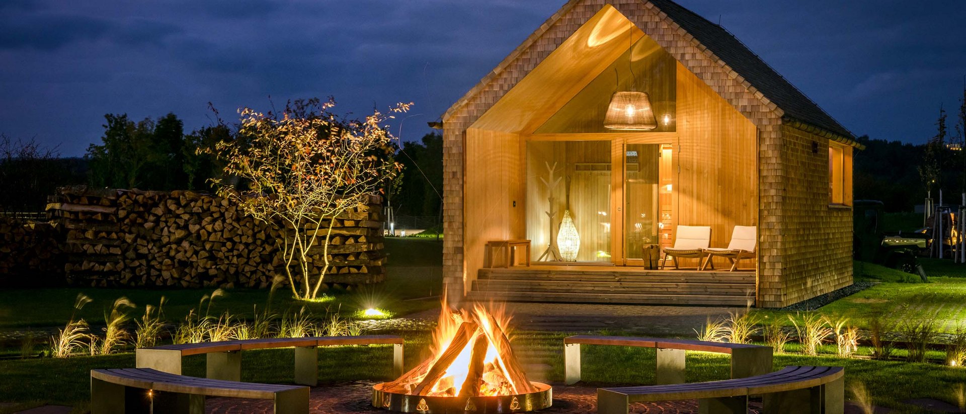 Seezeitlodge: Bildergalerie Holzhaus bei Nacht mit Lagerfeuer und Sitzbänken im Garten