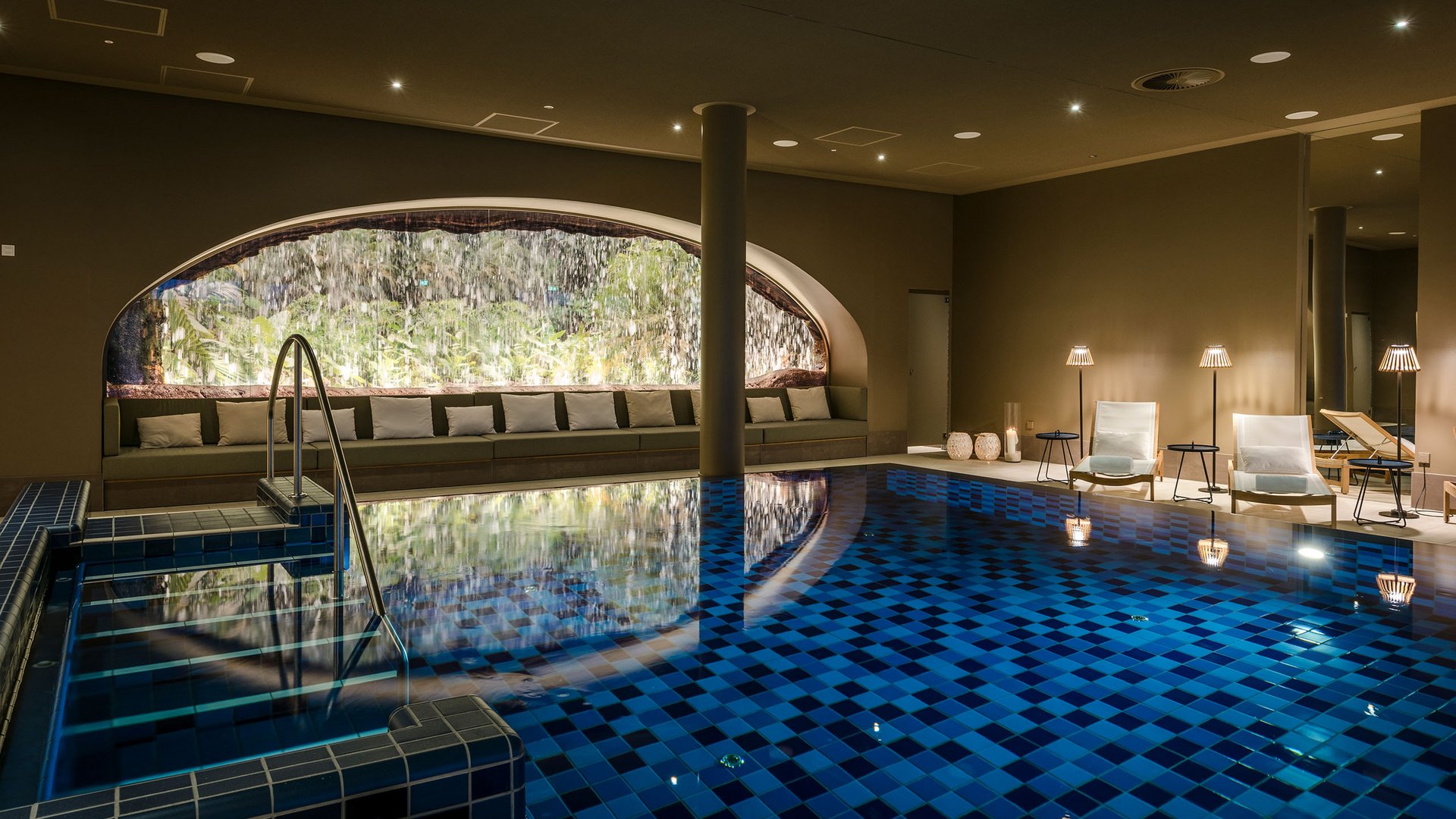Seezeitlodge: Bildergalerie Beheiztes Hallenbad mit Wasserfall und Loungemöbeln in warmem Licht
