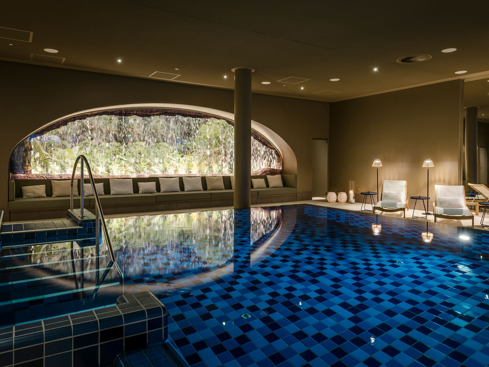 Seezeitlodge: Bildergalerie Beheiztes Hallenbad mit Wasserfall und Loungemöbeln in warmem Licht