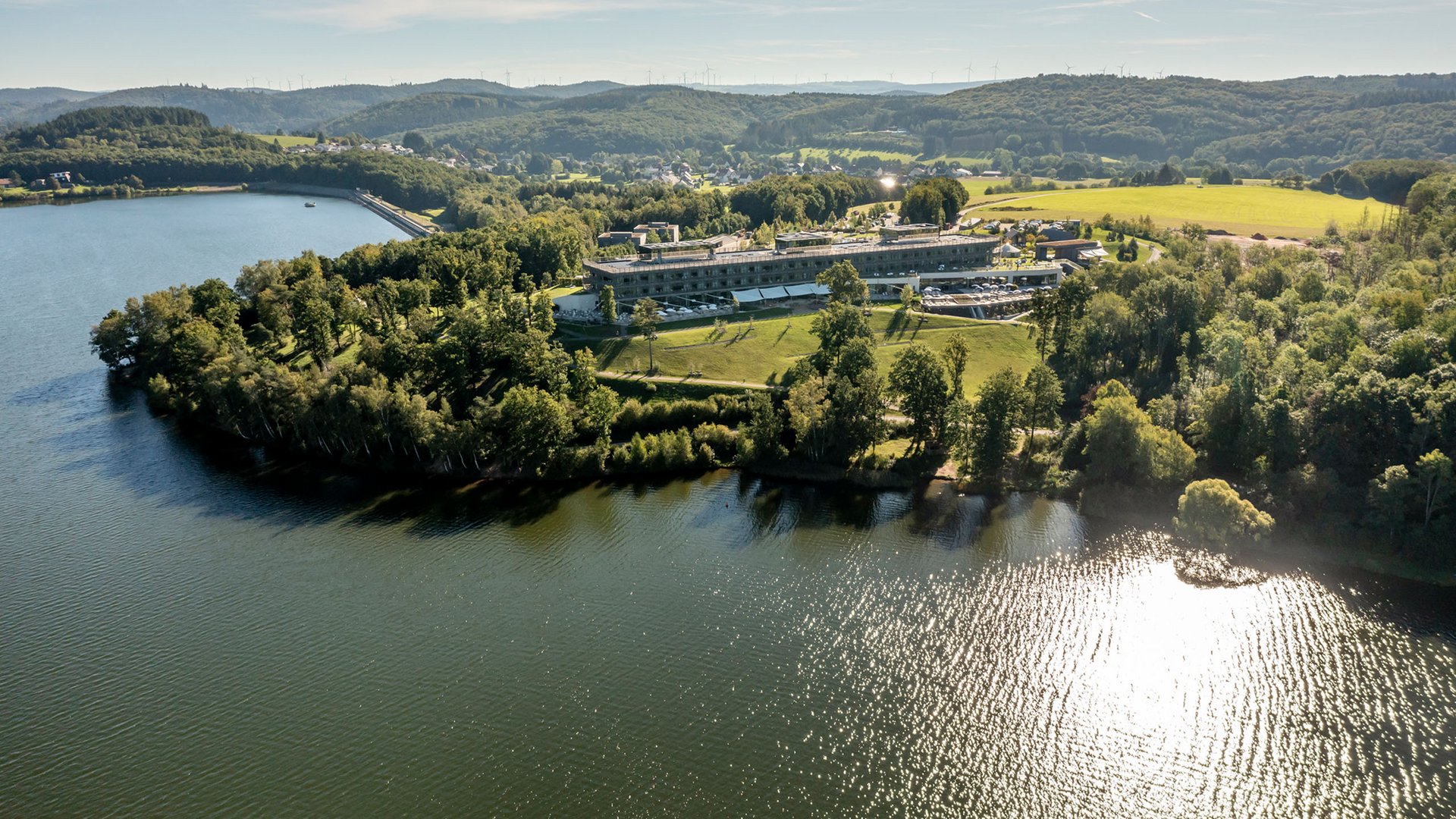 Seezeitlodge: Lieblingsgeschichten Luftaufnahme von einem See mit einem Gebäude und bewaldeten Hügeln im Hintergrund