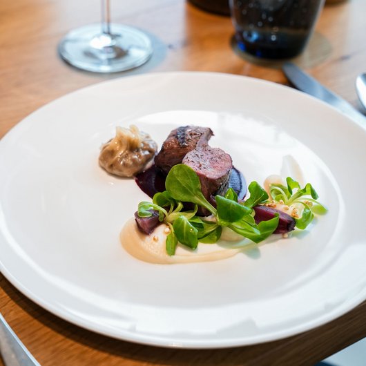 Ihr Genusshotel am Bostalsee Elegantes Gericht mit Fleisch, Sauce, grünem Salat und Rotwein im Hintergrund