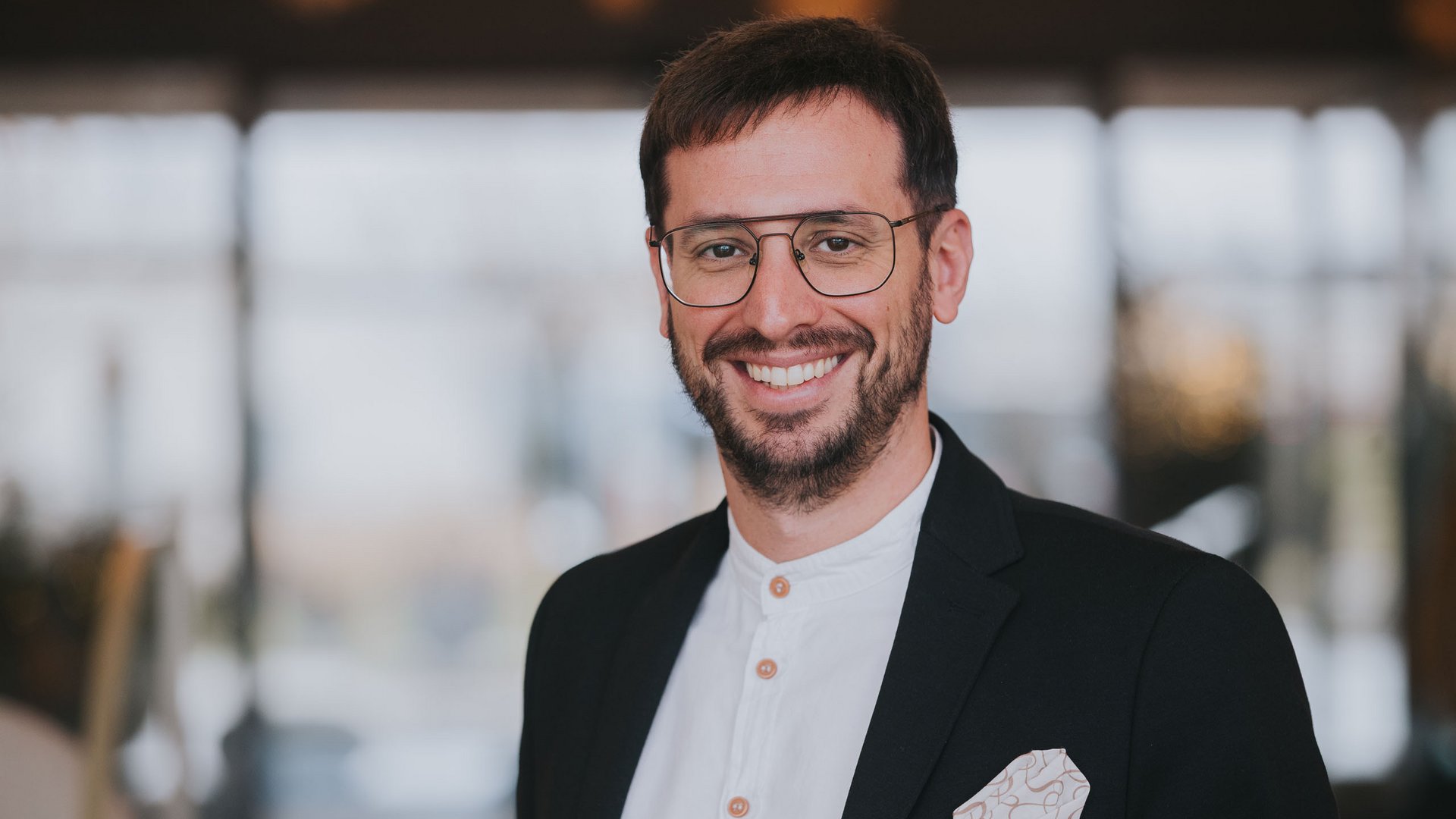 Seezeitlodge: Gastgeber mit Herz Lächelnder Mann mit Brille in Business-Outfit vor verschwommenem Hintergrund