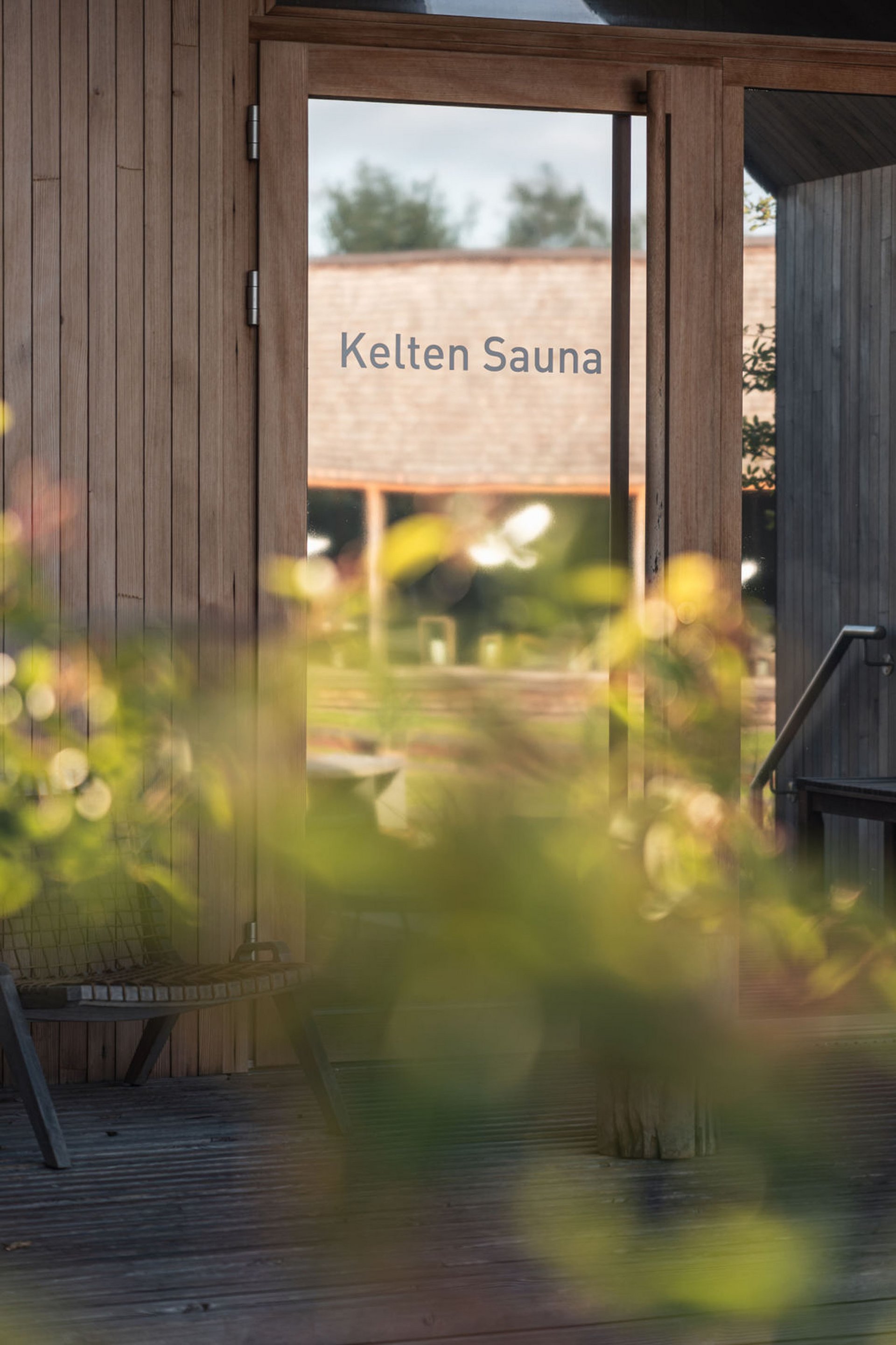 Seezeitlodge : Galerie photos Porte en verre avec inscription Kelten Sauna et mur en bois avec plantes floues au premier plan