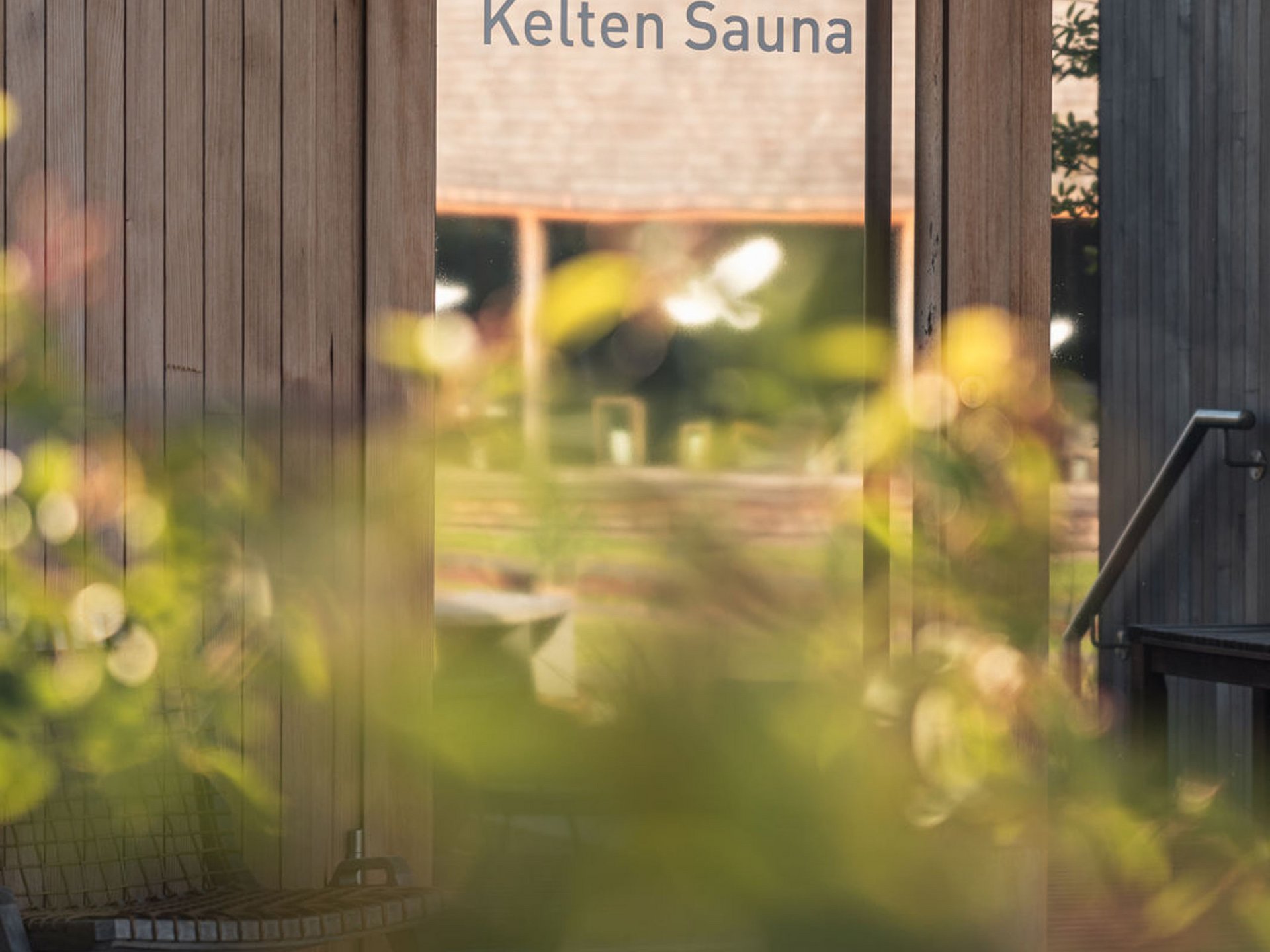 Seezeitlodge : Galerie photos Porte en verre avec inscription Kelten Sauna et mur en bois avec plantes floues au premier plan