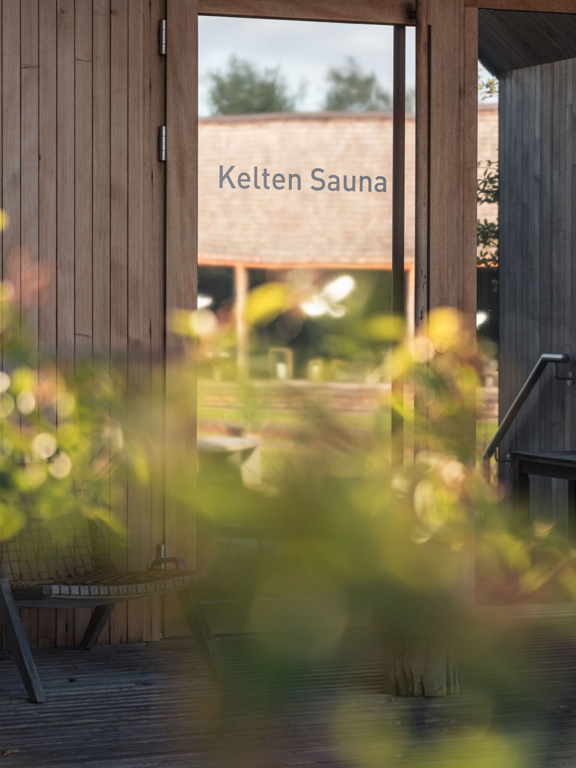 Seezeitlodge : Galerie photos Porte en verre avec inscription Kelten Sauna et mur en bois avec plantes floues au premier plan