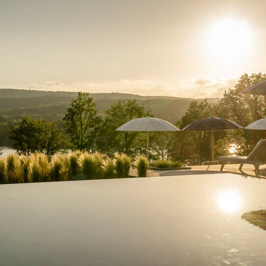 Le Seezeitlodge : votre hôtel au lac de Bostal Piscine avec chaises longues et parasols au coucher du soleil avec forêt en arrière-plan