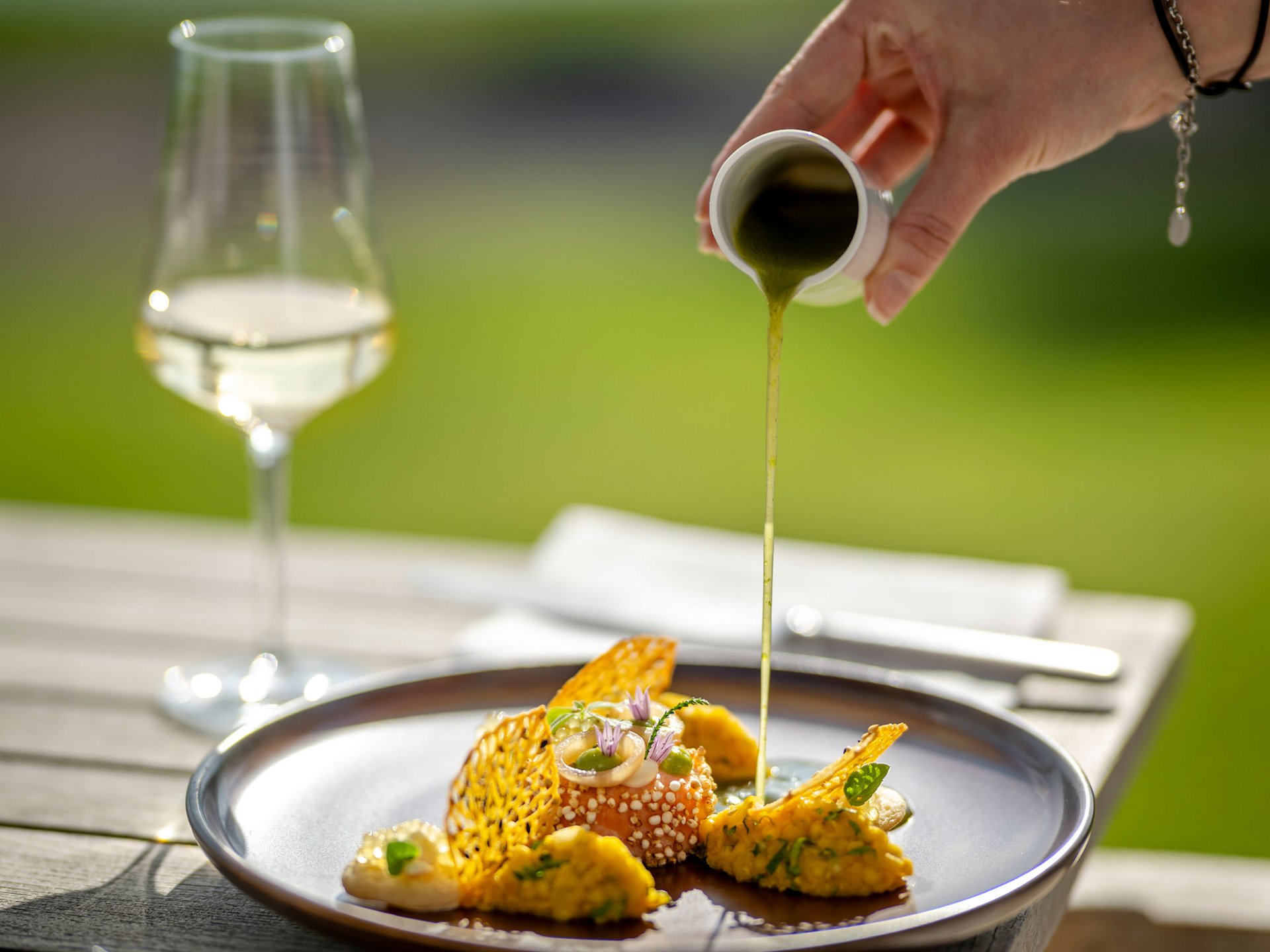 Seezeitlodge: Bildergalerie Hand gießt grüne Sauce über eine kunstvoll angerichtete Gourmet-Mahlzeit im Freien