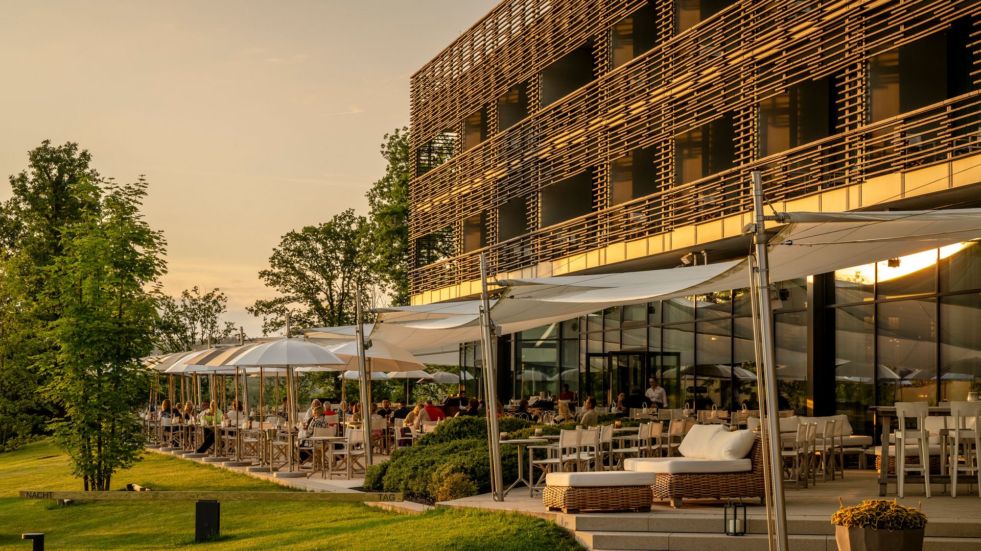Seezeitlodge: Bildergalerie Sonnenterrasse eines Restaurants mit Gästen und moderner Holzfassade bei Sonnenuntergang