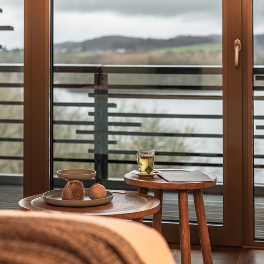 Übernachten am Bostalsee: Seezeitlodge Hotel & Spa Gemütliche Holzläche mit Blick auf See hinter Glas-Schiebetür