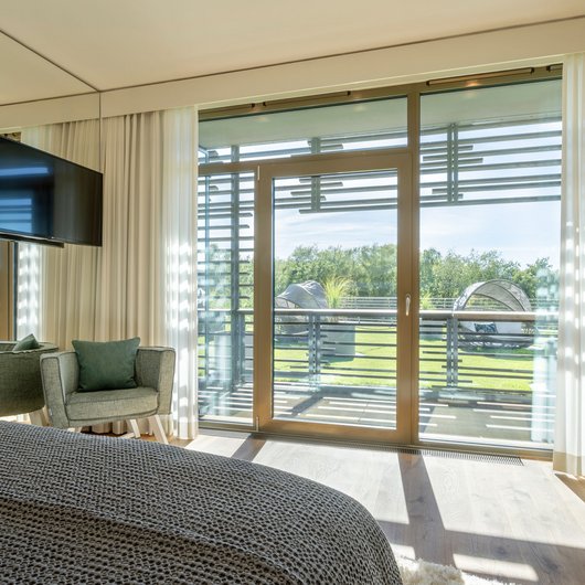 Übernachten am Bostalsee: Seezeitlodge Hotel & Spa Helles Schlafzimmer mit Sessel, TV und Balkon mit Aussicht auf Garten